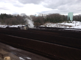 Rows of dark soil or compost piles in an outdoor facility with trees and light snow in the background.