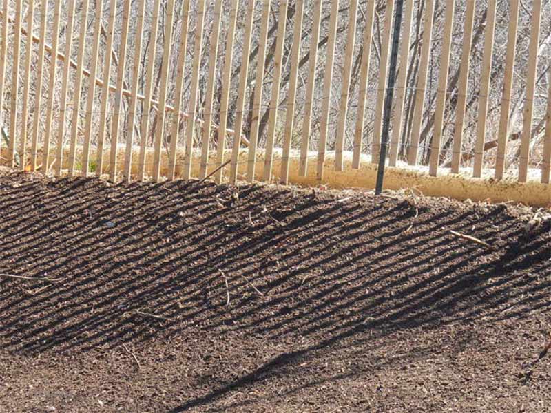 Wooden fence casting diagonal shadows across a freshly graded soil slope.