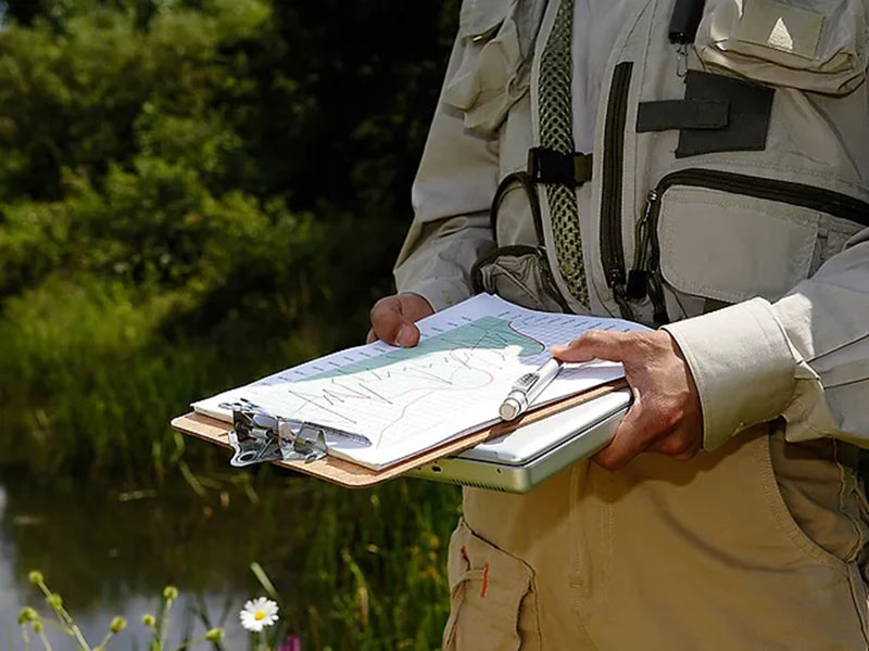 Person holding a clipboard and map near a grassy outdoor area.