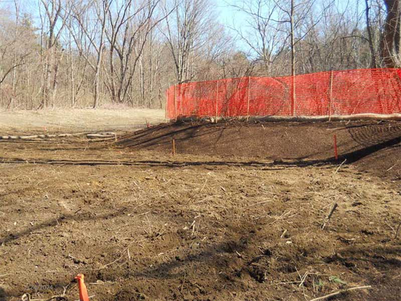 Freshly graded soil area with orange safety fencing along a slope and trees in the background.
