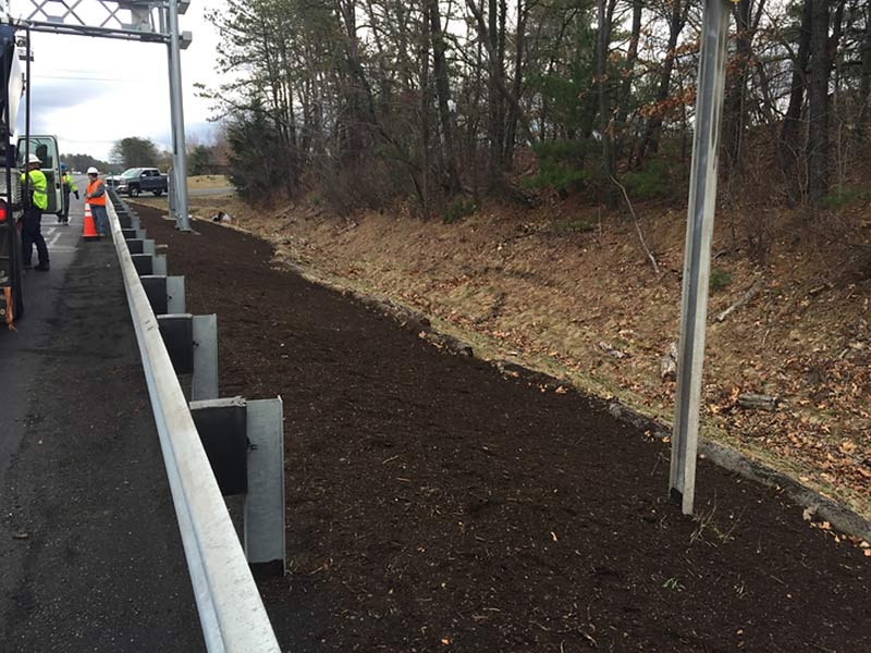 Freshly mulched roadside area beside a guardrail on a highway near a wooded slope.