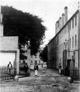 Historic black‑and‑white photo of people standing in a cobblestone alley between old buildings.