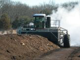 Large industrial machine working on a pile of soil with steam rising in the background.