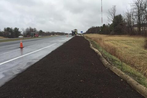Freshly mulched roadside area beside a wet highway on a cloudy day.