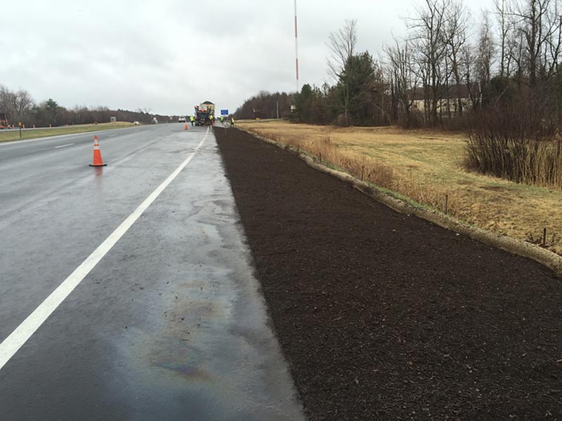 Freshly mulched roadside area next to a wet highway with traffic cones and maintenance vehicles ahead.