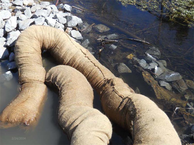 Sediment control booms floating in shallow water near rocks.