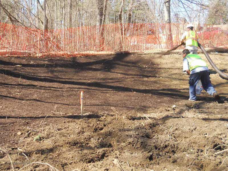 Workers using hoses on a soil-covered slope near an orange safety fence.