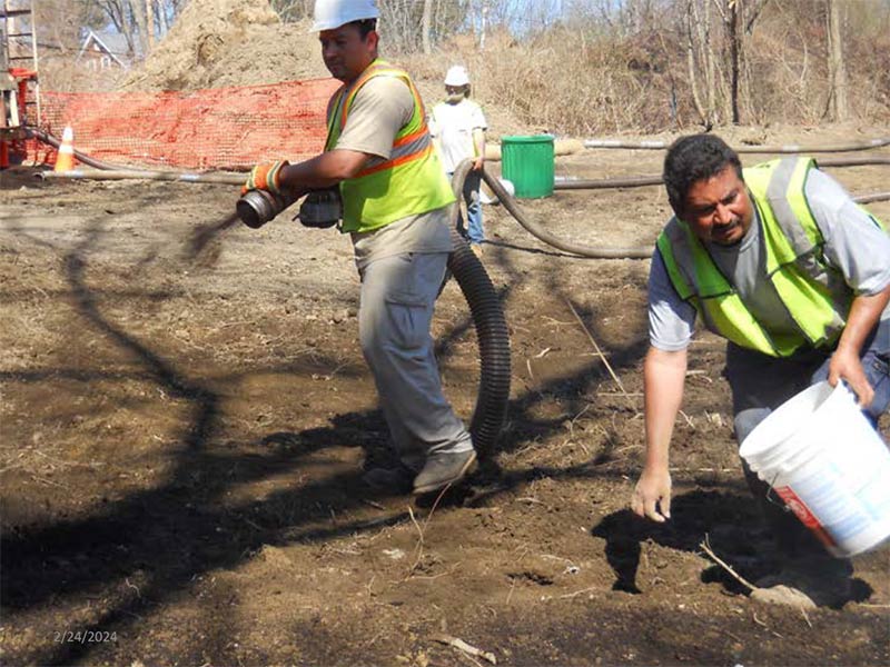 Workers performing tasks on a dirt worksite with hoses and a bucket, near an orange safety fence.